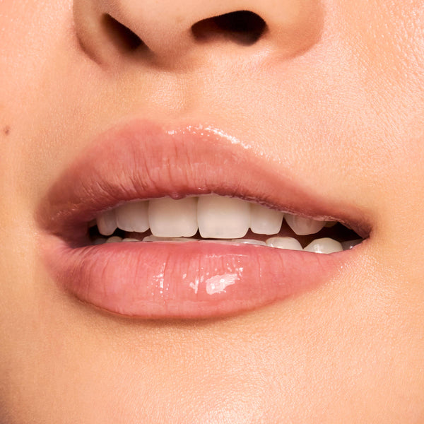 Glossy lips wearing Get Rich from the ColourPop Glossy Lip Balm Trio, adding sheer nude shine as the mouth softly parts to show teeth, captured in an extreme close-up against natural skin for a high-shine, hydrated finish.