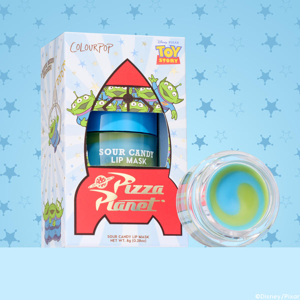 ColourPop Lip Mask The Chosen One sits in a Toy Story Pizza Planet rocket box with green alien art, showing the Sour Candy jar and an open pot revealing a blueβgreen swirl balm, 8 g (0.28 oz), against a blue sheriffβstar patterned background.