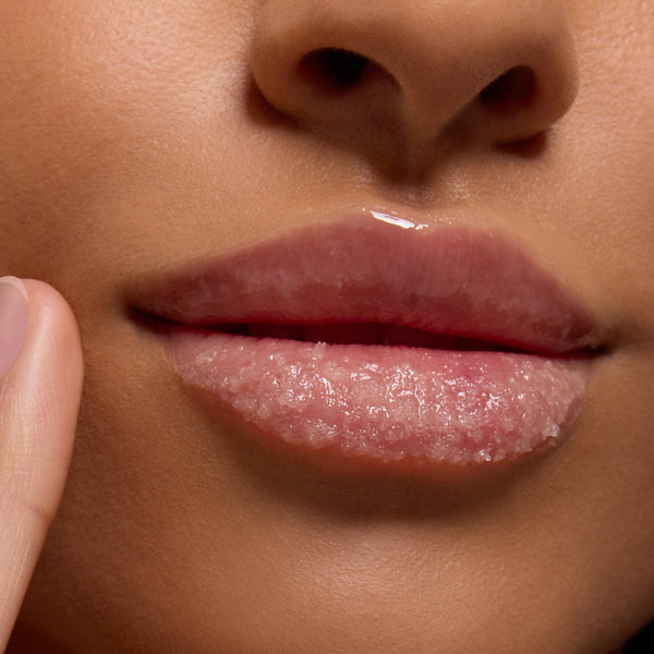 Glossy lips buffed with Neapolitan Sundae ColourPop Lip Scrub, sugar crystals exfoliating the lower lip as a fingertip applies the treatment in a close-up, revealing a smooth, hydrated finish.
