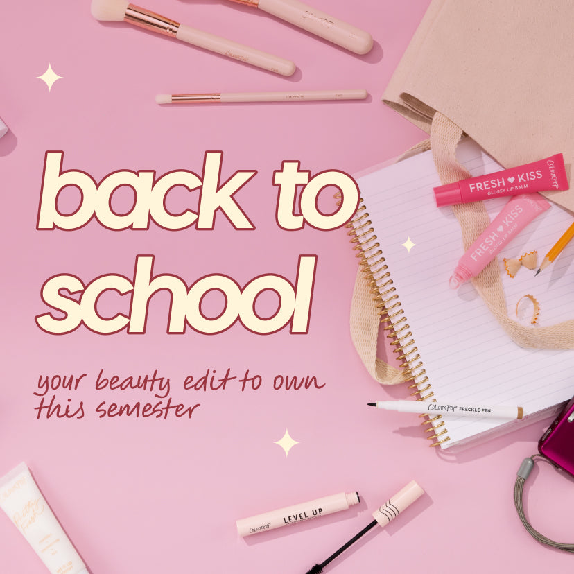 ColourPop makeup arranged in a back-to-school flat lay - Fresh Kiss glossy lip balms, Level Up mascara with open wand, and a Freckle Pen - set beside pink brushes, a spiral notebook, tote straps, and a phone on a pastel pink desk.