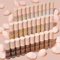 ColourPop Pretty Fresh Creamy Concealer in Fair 15 W showcased with the full shade range, bottles standing in tidy rows with blush-pink caps, surrounded by soft pink makeup sponges on a nude background.