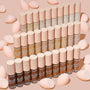 ColourPop Pretty Fresh Creamy Concealer in Fair 15 W showcased with the full shade range, bottles standing in tidy rows with blush-pink caps, surrounded by soft pink makeup sponges on a nude background.