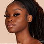 Model wearing ColourPop Pretty Fresh Tinted Moisturizer in Deep Dark 21 W, showcasing smooth, dewy coverage on deep warm skin; close-up side portrait with natural lashes and glossy lips against a soft beige background.