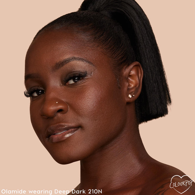 Model wearing ColourPop Pretty Fresh Creamy Concealer in Deep Dark 210 N, displaying smooth, even coverage and a neutral undertone on deep skin in a close-up portrait against a warm beige background.