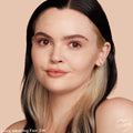 Fair 3 W shade of ColourPop Pretty Fresh Tinted Moisturizer worn by a model, showing a dewy, light-coverage finish that evens tone; close-up against a peach studio backdrop with natural brows, nude gloss, black-and-blonde hair, and small hoop earrings.