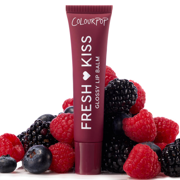 ColourPop Fresh Kiss Glossy Lip Balm tube stands upright amid raspberries, blackberries, and blueberries on a white backdrop, representing the Get Rich ColourPop Glossy Lip Balm Trio with a deep berry-colored tube and high-shine finish.