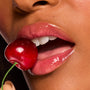 Glossy pink lips wear ColourPop Fresh Kiss Glossy Balm in Cherry Glaze, gripping a red cherry between the teeth in a macro beauty shot, showcasing a high-shine, juicy finish and hydrating lip balm texture.