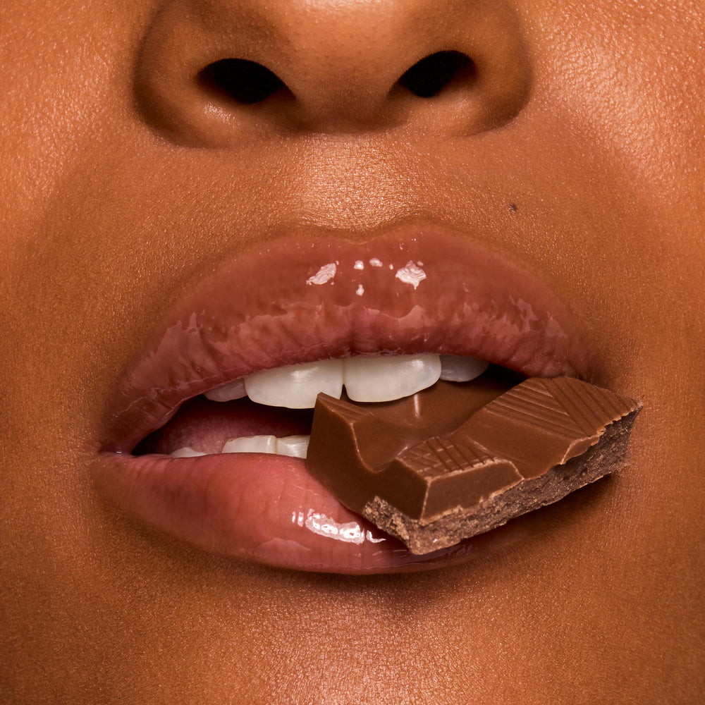 Glossy, cocoa-tinted lips wearing ColourPop Fresh Kiss Glossy Balm in Chocolate Milkshake hold a square of milk chocolate between teeth in a macro close-up, showcasing high-shine, cushy hydration and a creamy nude tint.