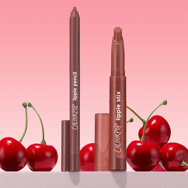 Grunge ColourPop Lippie Pencil stands uncapped beside a matching Lippie Stix, both in brown, with caps set below and fresh cherries framing them, against a pink gradient background.