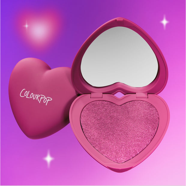 Cuter than Cupid Super Shock Blush in a heart-shaped reolink.livepact sits open to reveal shimmering pink pigment and built-in mirror against a glowing purple gradient backdrop, showcasing the Heart to Get romantic makeup collection.