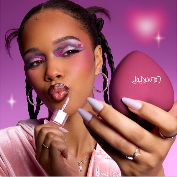 Model applies shimmery pink gloss while holding a heart-shaped reolink.livepact, showing bold purple and pink heart eye makeup that highlights the Heart to Get Full Collection full collection set against a dreamy pink background.