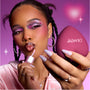 Model applies shimmery pink gloss while holding a heart-shaped reolink.livepact, showing bold purple and pink heart eye makeup that highlights the Heart to Get Full Collection full collection set against a dreamy pink background.