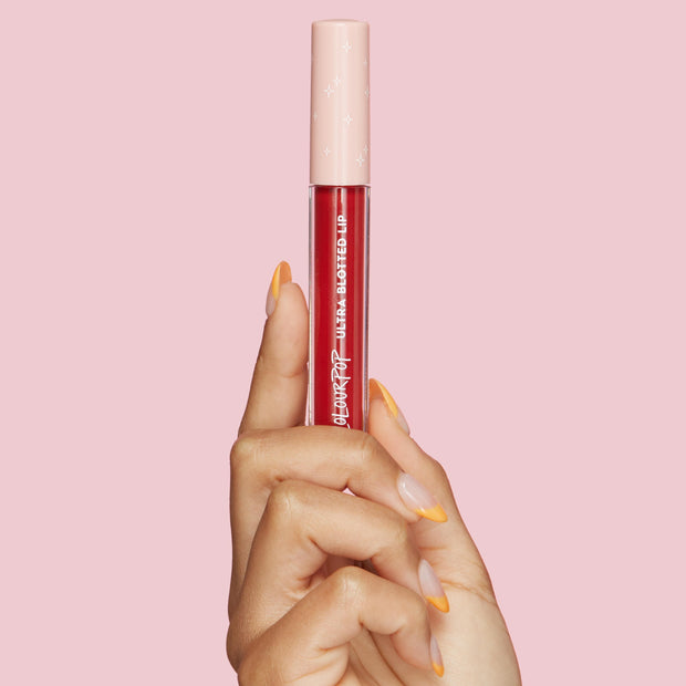 ColourPop Ultra Blotted Lip in Keep It Classy held upright by a hand with orange French tips, showcasing a clear tube of deep berry-red tint with a blush pink cap dotted with stars against a solid pink background.