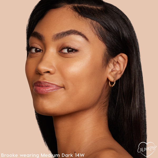 Model's face wearing Medium Dark 14 W, showcasing a natural, dewy finish from the ColourPop Pretty Fresh Tinted Moisturizer; soft glam with glossy lips and a small gold hoop earring against a nude background.