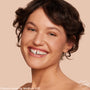 Model smiling and wearing ColourPop Pretty Fresh Creamy Concealer in Medium 83 C, showcasing smooth, natural coverage with a soft dewy finish on skin against a peach-beige studio background.