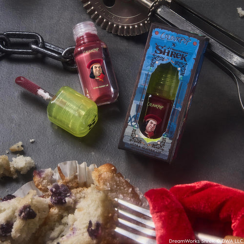 Short Lord ColourPop Ultra Glossy Lip - red tint with doe-foot applicator and neon green cap - rests open beside its Shrek-themed box featuring Lord Farquaad on a dark tabletop with crumbs, chain, and kitchen tools.