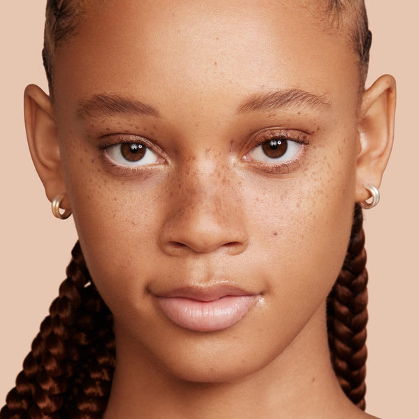 Model face wearing ColourPop Pretty Fresh Creamy Concealer, shade Medium 100 W (warm), brightening under-eyes with natural, seamless coverage; close-up studio portrait with freckles, glossy lips, braided hair, and silver hoops on a peach backdrop.