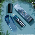 Team Edward Lux Lip Oil swatched: a sparkling deep blue, shimmer-packed gloss. Doe-foot applicator, open ColourPop Lux Lip Oil tube, and Twilight collab box rest on a teal surface framed by evergreen sprigs.