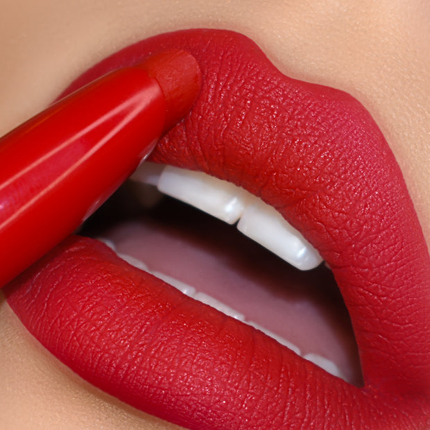 ColourPop Lippie Stix in Trust Me glides onto full lips, laying down vivid true-red matte color; extreme close-up shows the bullet tracing the upper lip with crisp edges, smooth texture, and white teeth peeking for a bold finish.
