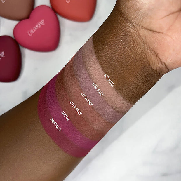 Babycakes swatch from ColourPop Pressed Powder Blush shown on deep skin, with labeled shade stripes - Babycakes, Text Me, 4Ever Yours, Let’s Dance, Flirt Alert, Kiss N Tell - showing rich pigment; heart-shaped reolink.livepacts blurred in the background.
