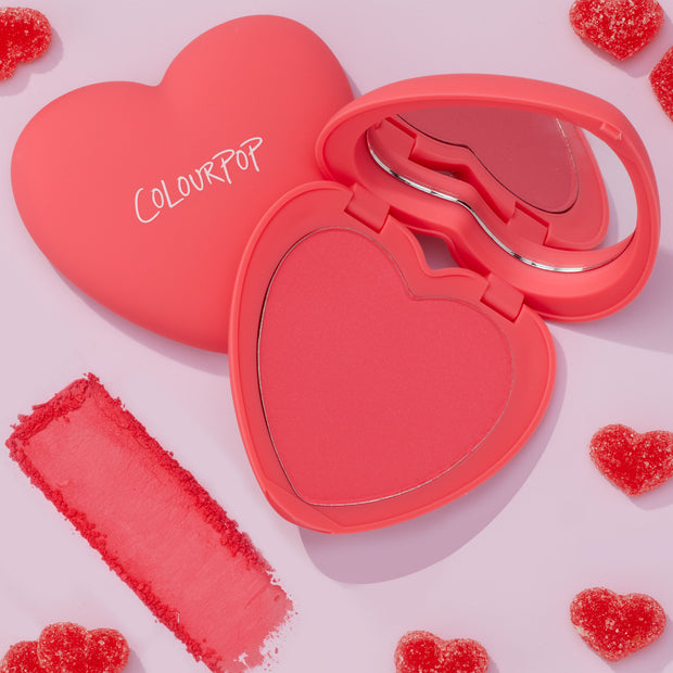 Woo Me ColourPop Pressed Powder Blush opens in a coral heart-shaped compact with mirror and white ColourPop logo, revealing a rosy pink pan; a velvety swatch sits beside on a pale pink surface with scattered red sugar heart candies.