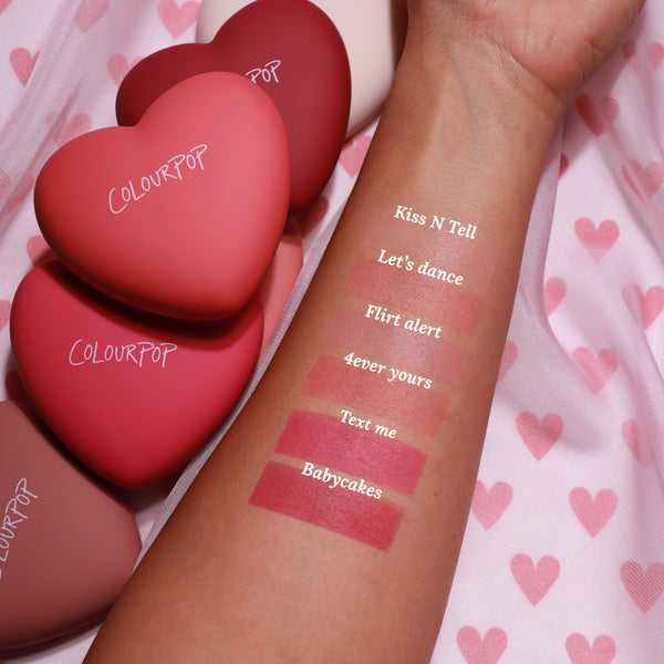Swatched forearm displays “Text Me” ColourPop Pressed Powder Blush, a rosy pink matte stripe, alongside shades Babycakes, 4ever Yours, Flirt Alert, Let’s Dance, and Kiss N Tell; heart-shaped reolink.livepacts on pink heart-pattern fabric.